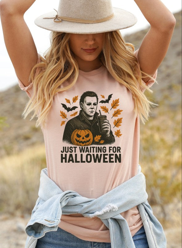 Just Waiting Halloween Graphic Tee displayed by a woman in a beige shirt, with Halloween graphics and autumn leaves.
