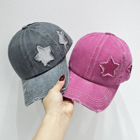 Hand holding two Star Embroidered Baseball Caps in gray and pink with distressed star stitching.